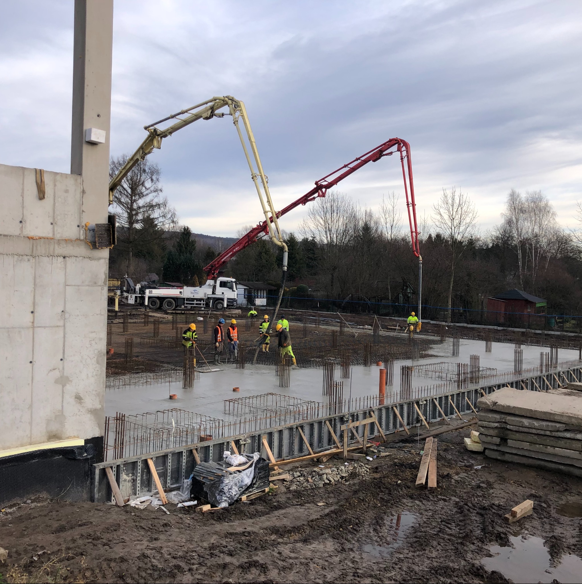 Wylewanie betonu na fundamenty zbrojone. Widoczne pompy do betonu, pracownicy w kaskach i kamizelkach odblaskowych. Szalunki i wystające pręty zbrojeniowe.