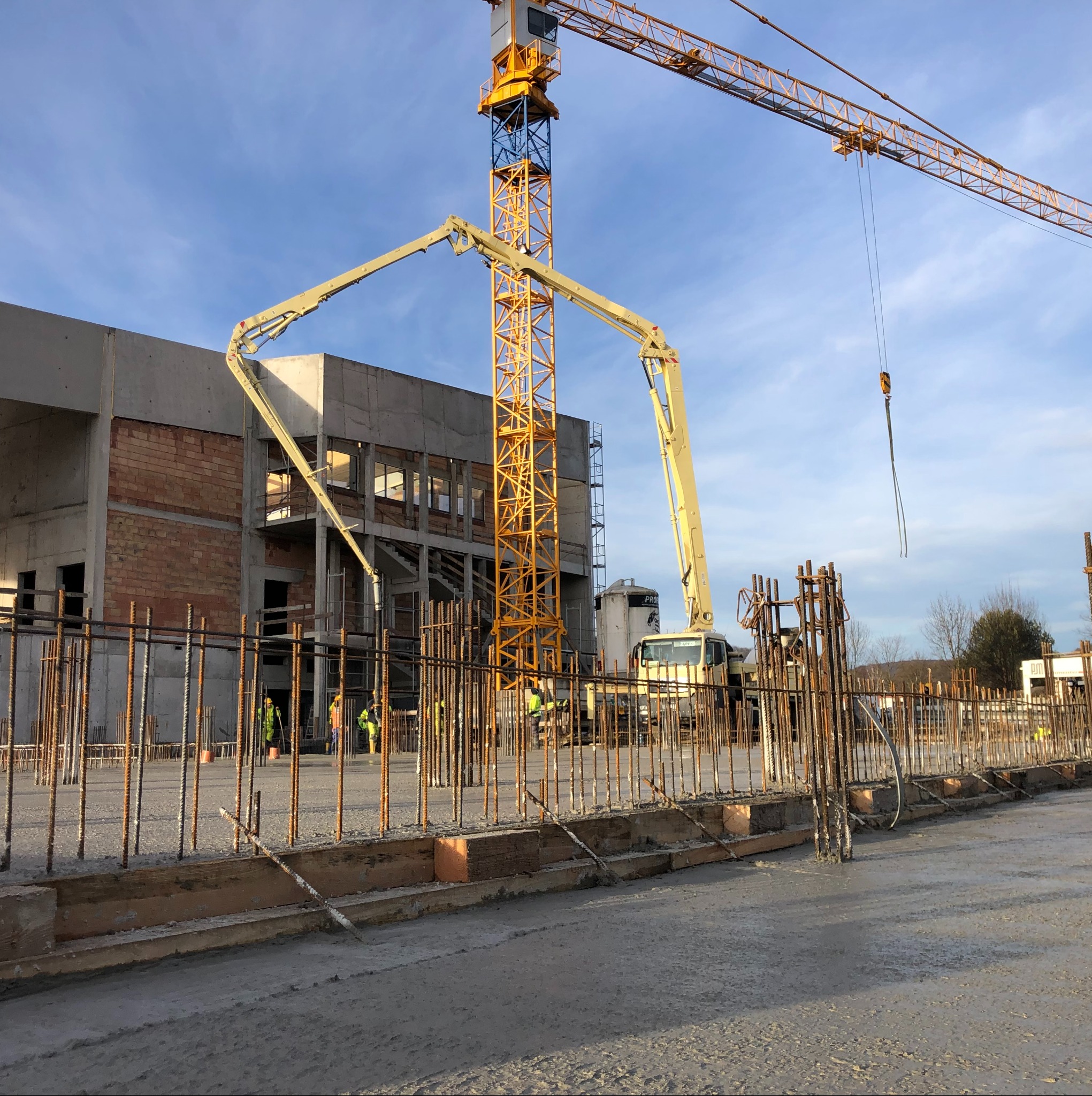 Wylewanie betonu na zbrojony fundament budynku w trakcie budowy. Żuraw budowlany i pracownicy w tle. Widok na plac budowy w słoneczny dzień.