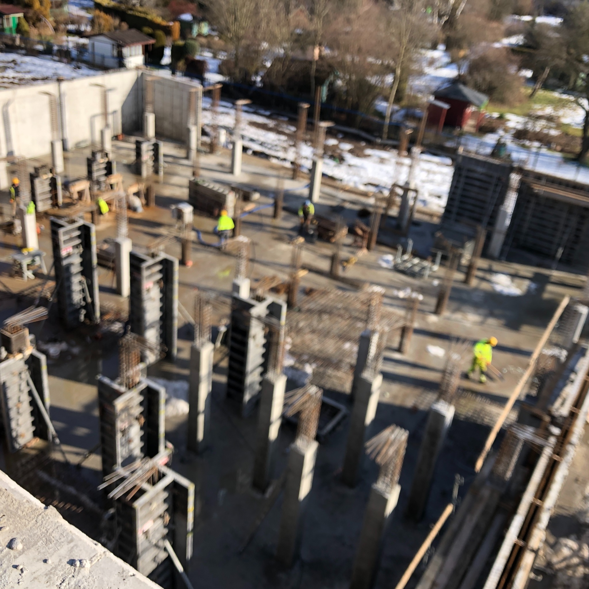 Budowa budynku w trakcie: widoczne zbrojenia, szalunki i pracownicy w żółtych kamizelkach. Ujęcie z góry, w tle zabudowa jednorodzinna i drzewa.