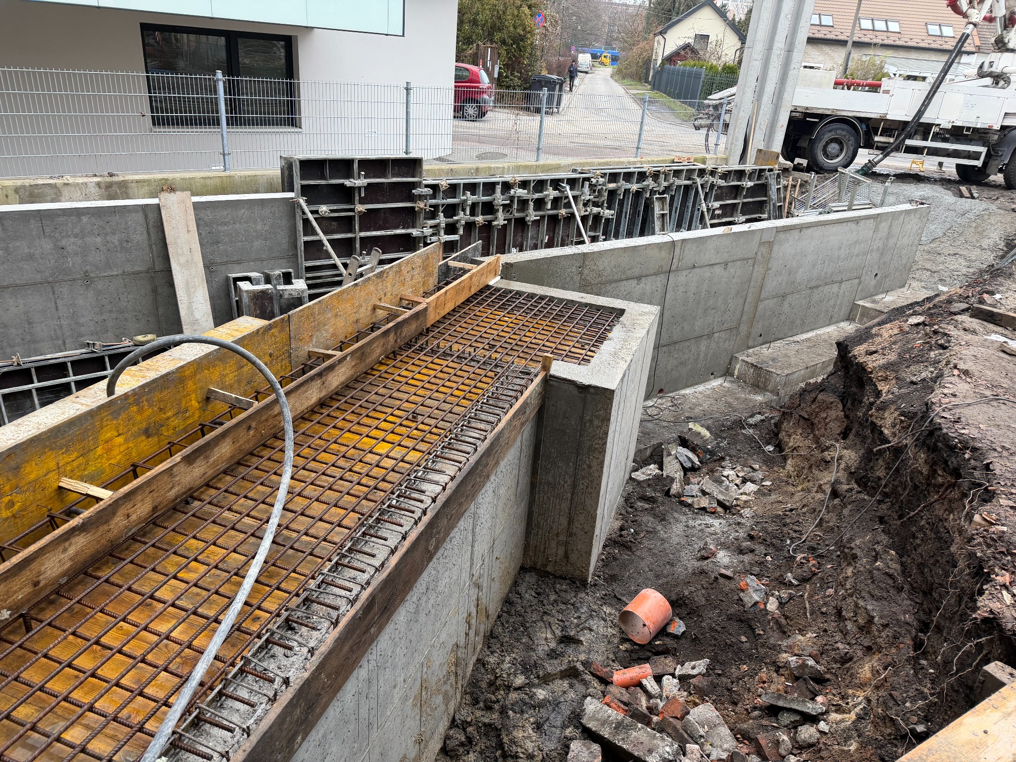 Zbrojenie i szalunek fundamentów budynku w trakcie budowy. Widoczne betonowe ściany fundamentowe, stalowe pręty zbrojeniowe i drewniane elementy szalunku.