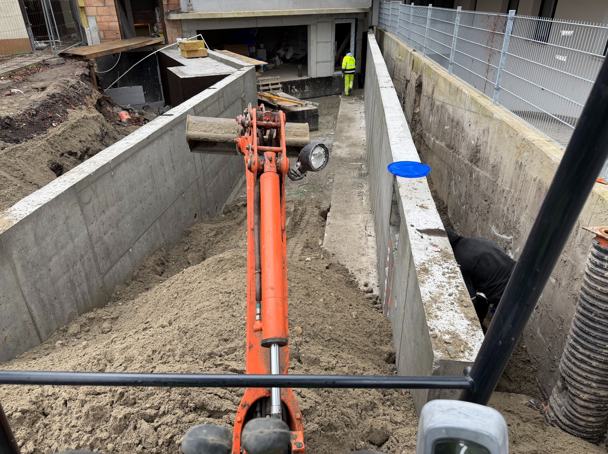 Mini koparka w wykopie fundamentowym z betonowymi ścianami, widok z kabiny operatora. W tle pracownik w odblaskowej kamizelce.