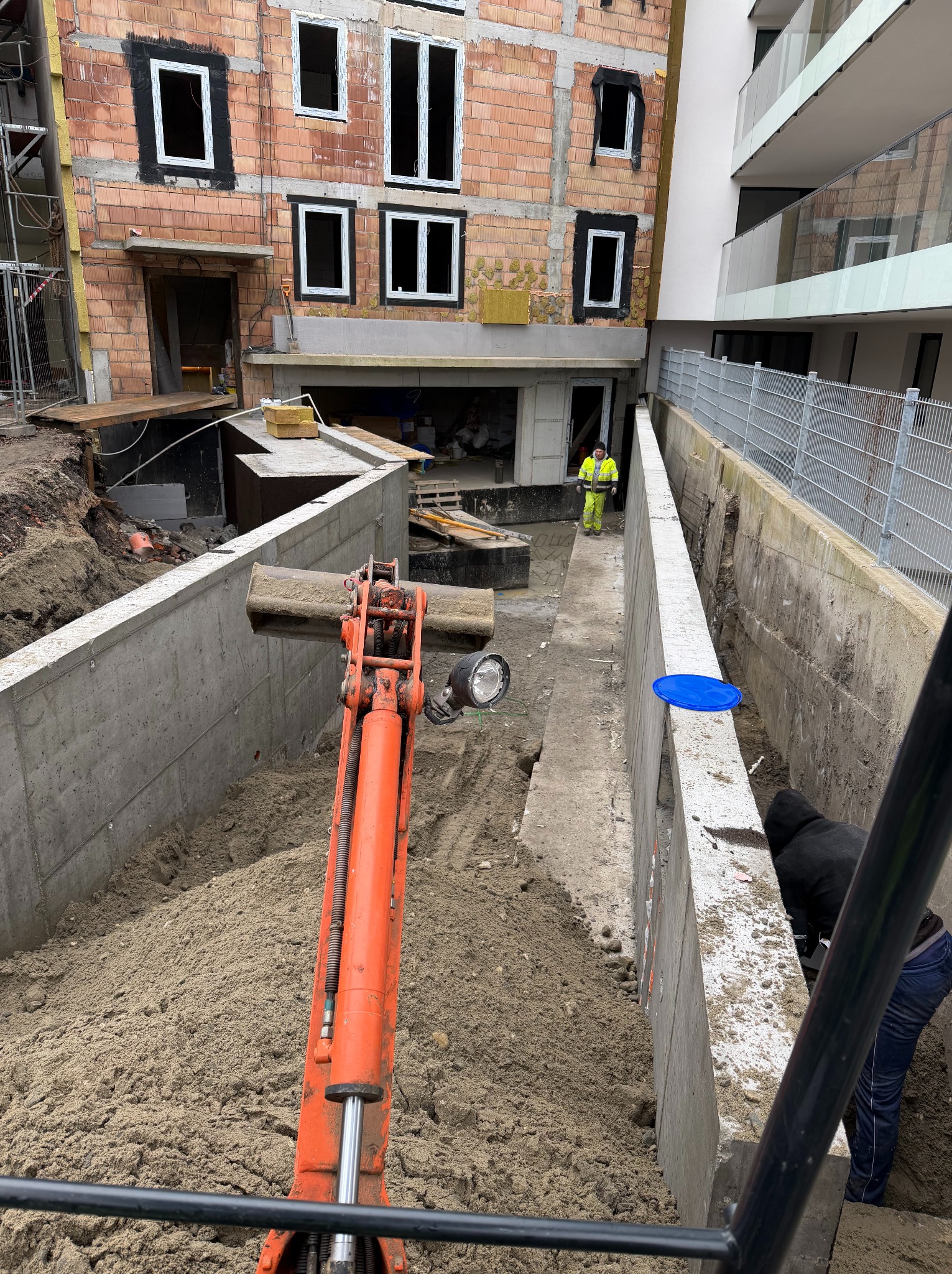 Wykop pod fundamenty budynku wielorodzinnego z użyciem pomarańczowej koparki. Widoczny pracownik w żółtej kamizelce i fragment budynku w stanie surowym.