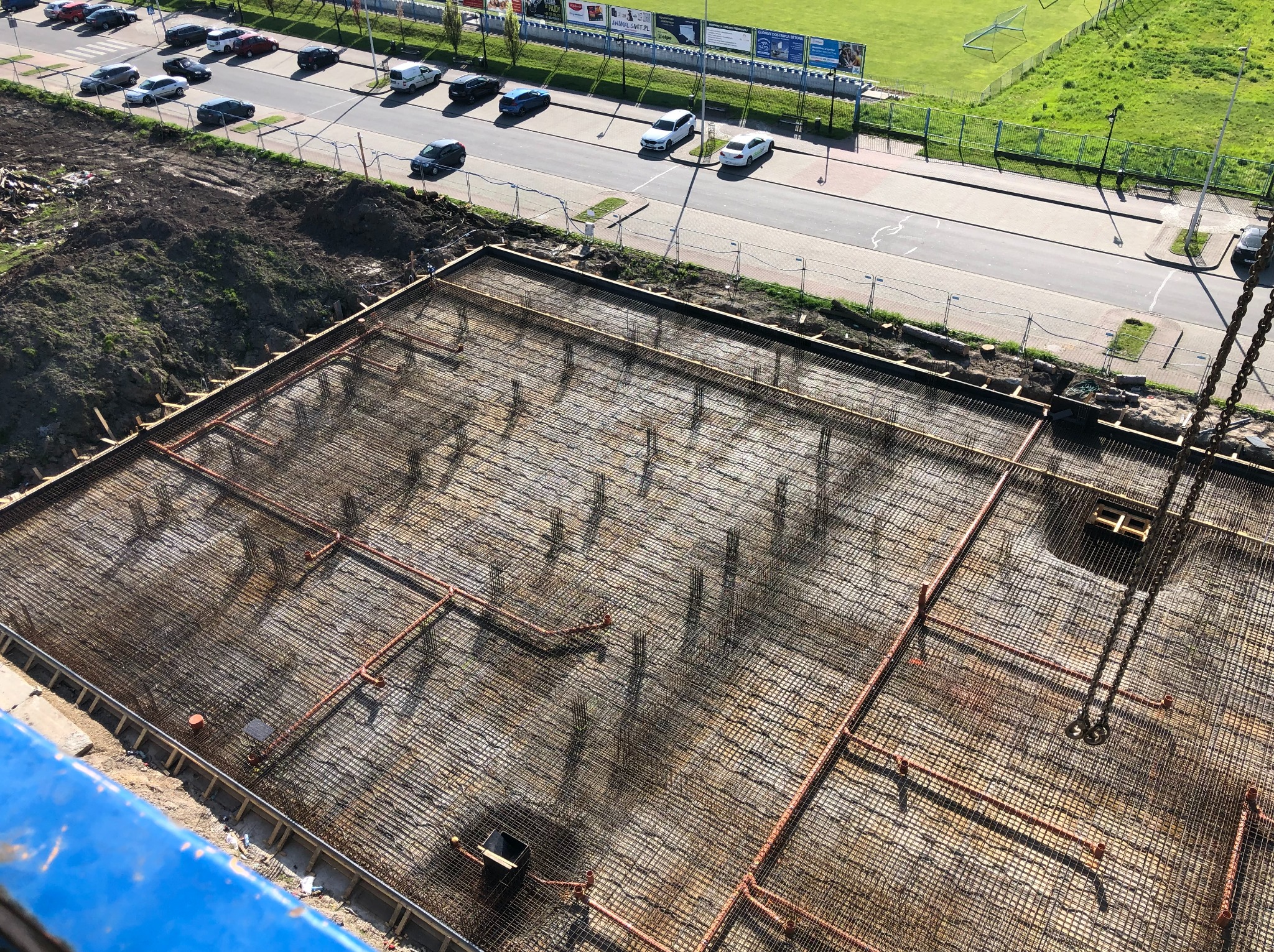 Zbrojenie fundamentu z wystającymi prętami i rurami instalacyjnymi, widok z góry na plac budowy z ulicą i zielenią w tle.