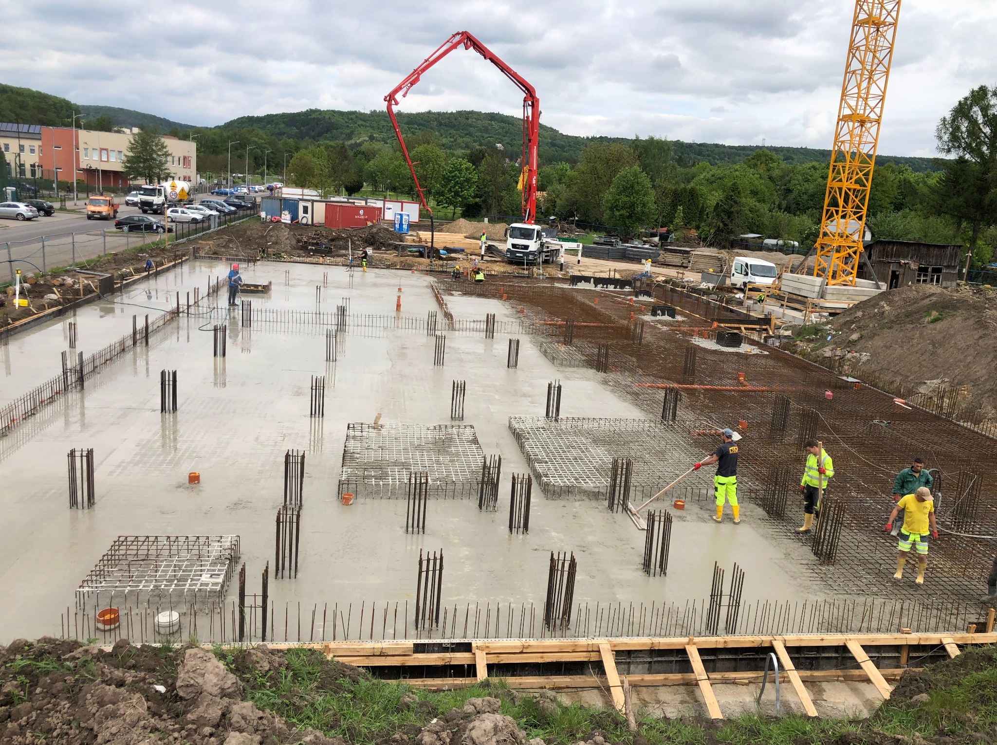 Wylany fundament z wystającymi prętami zbrojeniowymi, ekipa budowlana wygładza beton, w tle pompa do betonu i żuraw wieżowy, pochmurne niebo.