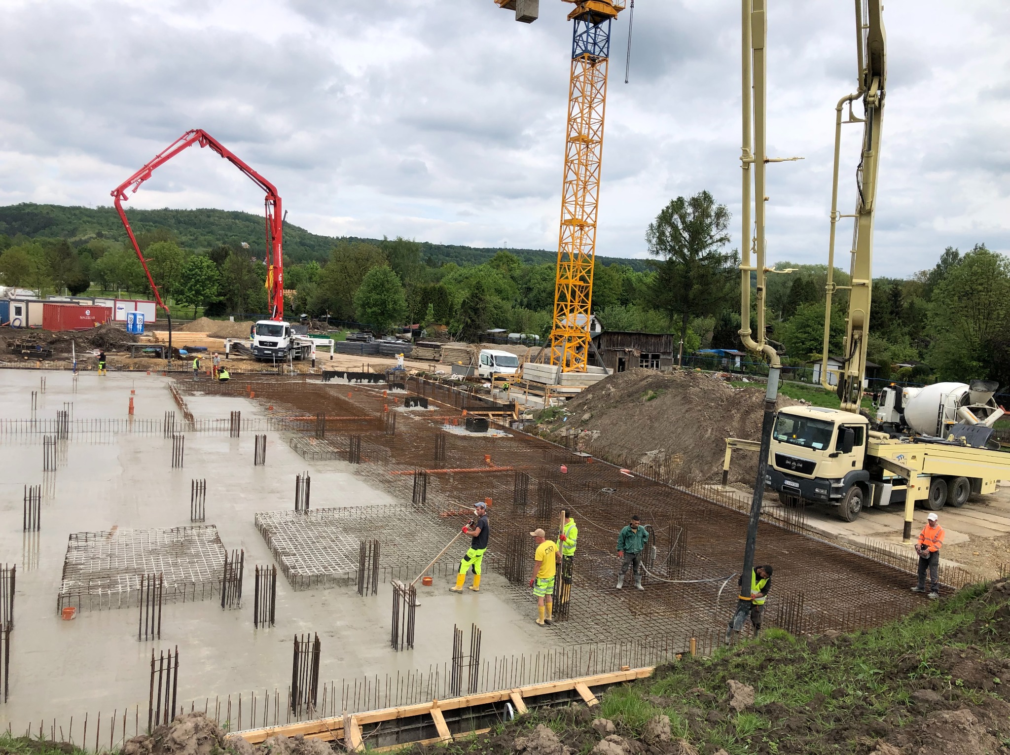 Wylewanie betonu na zbrojone fundamenty budynku. Na budowie pracownicy, pompy do betonu i żurawie. W tle zieleń i pochmurne niebo.