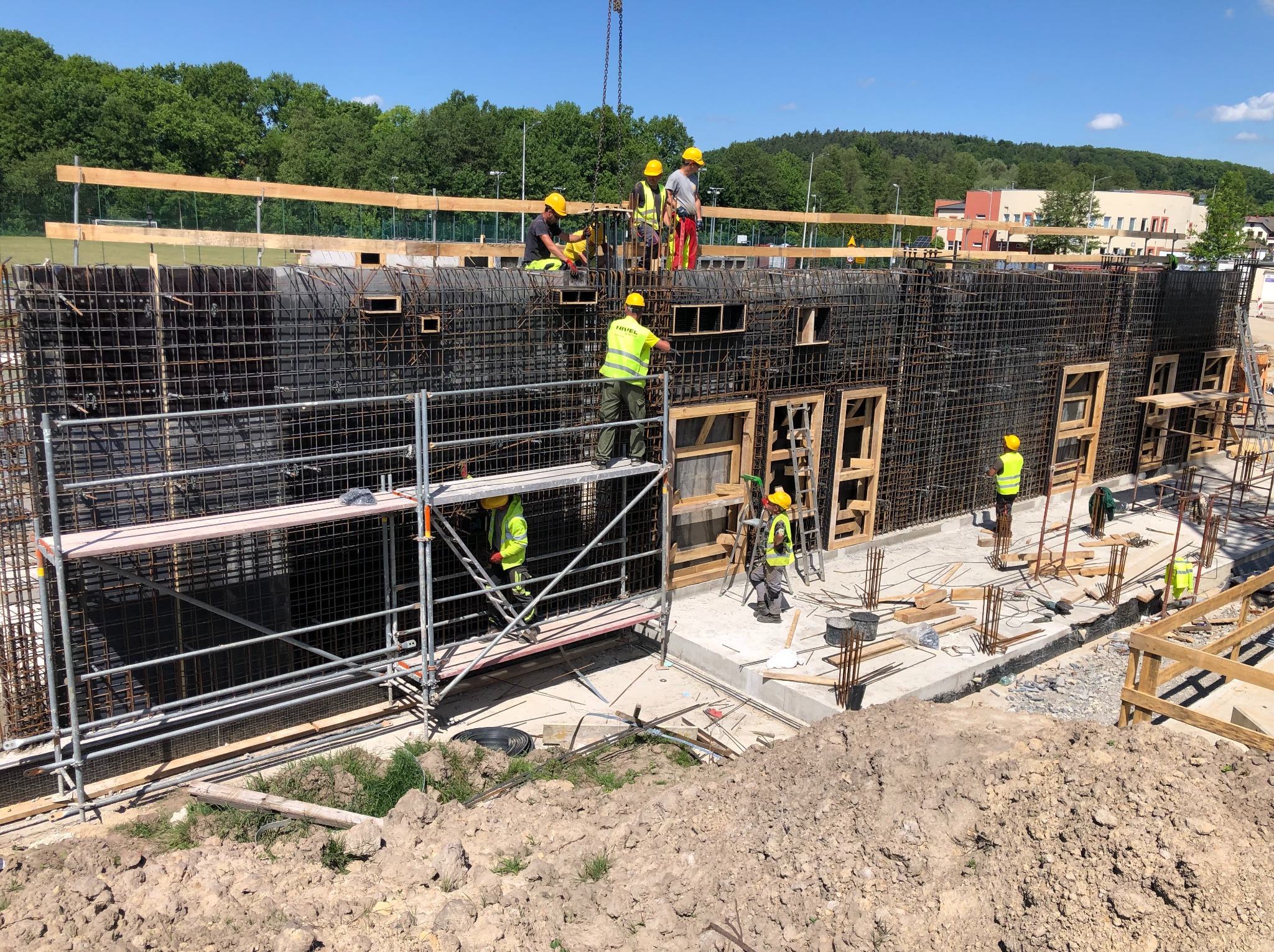 Budowa budynku żelbetowego: zbrojenie ścian z otworami okiennymi, pracownicy w kaskach na rusztowaniach, dźwig podnosi element konstrukcyjny. Słoneczny dzień.