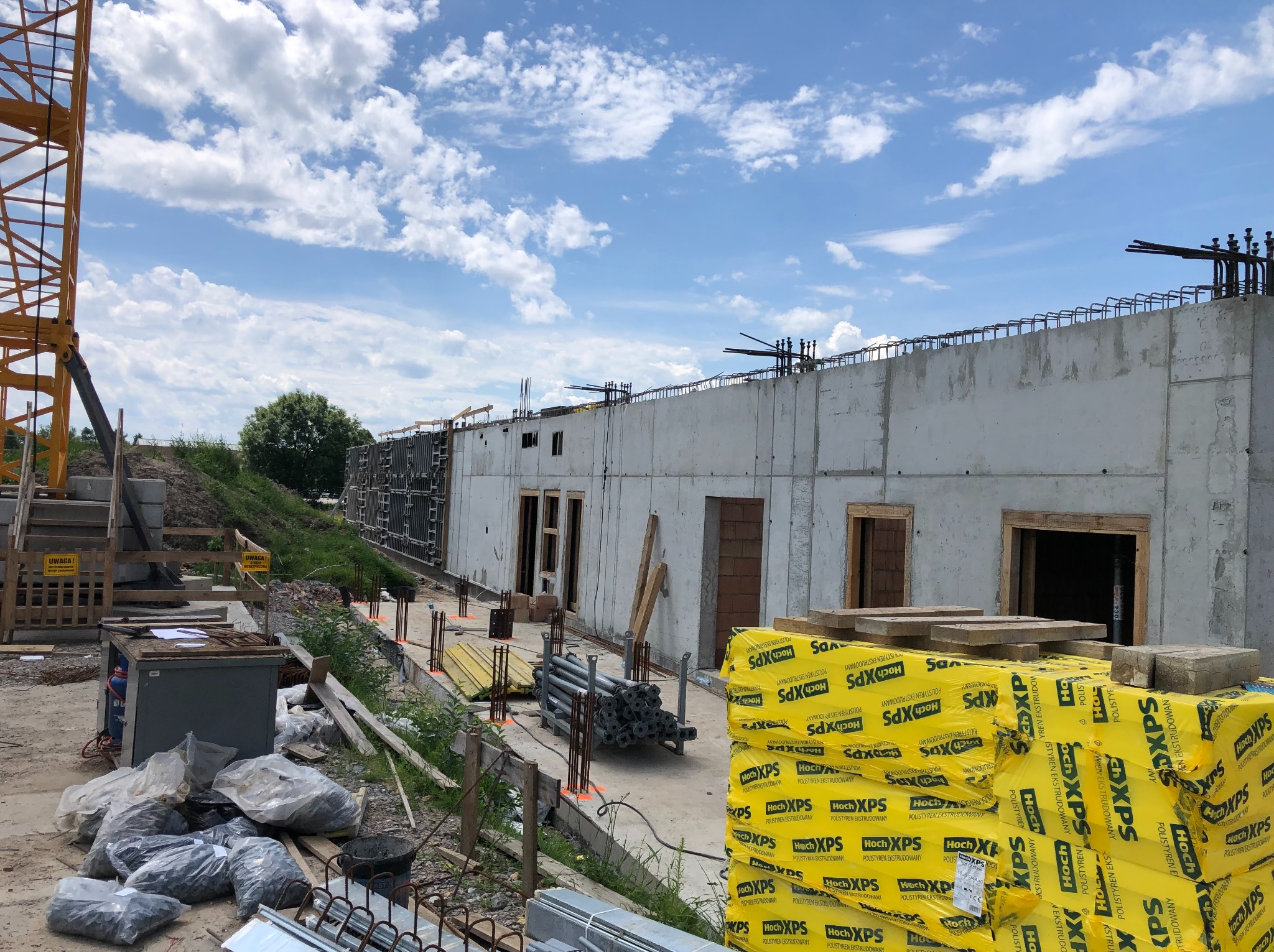 Betonowa konstrukcja budynku w trakcie budowy, widoczne zbrojenia i szalunki. Na pierwszym planie żółte paczki styropianu i materiały budowlane. W tle żuraw budowlany.