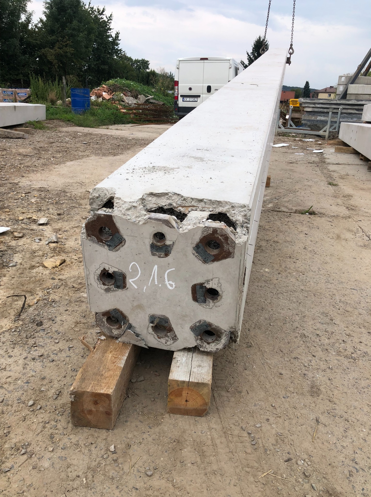 Prefabrykowany, betonowy element konstrukcyjny z widocznymi otworami i oznaczeniem '2,16', podwieszony na łańcuchu, oparty na drewnianych belkach na placu budowy.