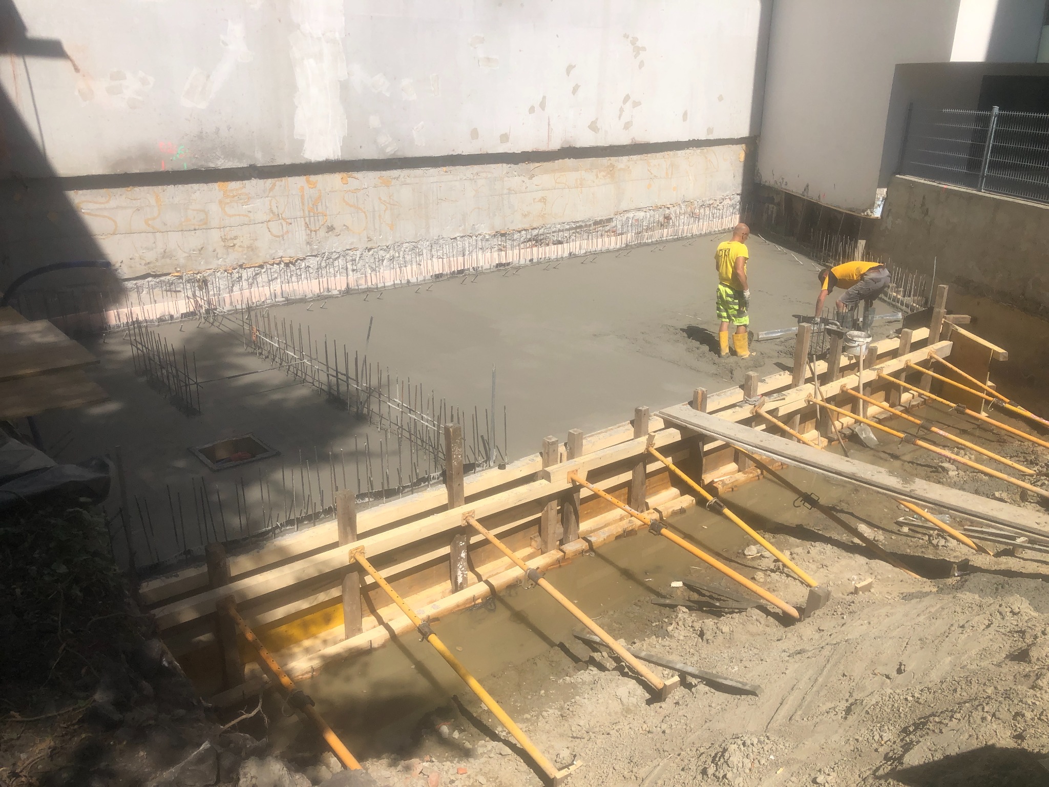 Wylany fundament zbrojony stalą, widoczne drewniane szalunki i dwóch pracowników w żółtych koszulkach podczas pracy. Ściany wykopu zabezpieczone.