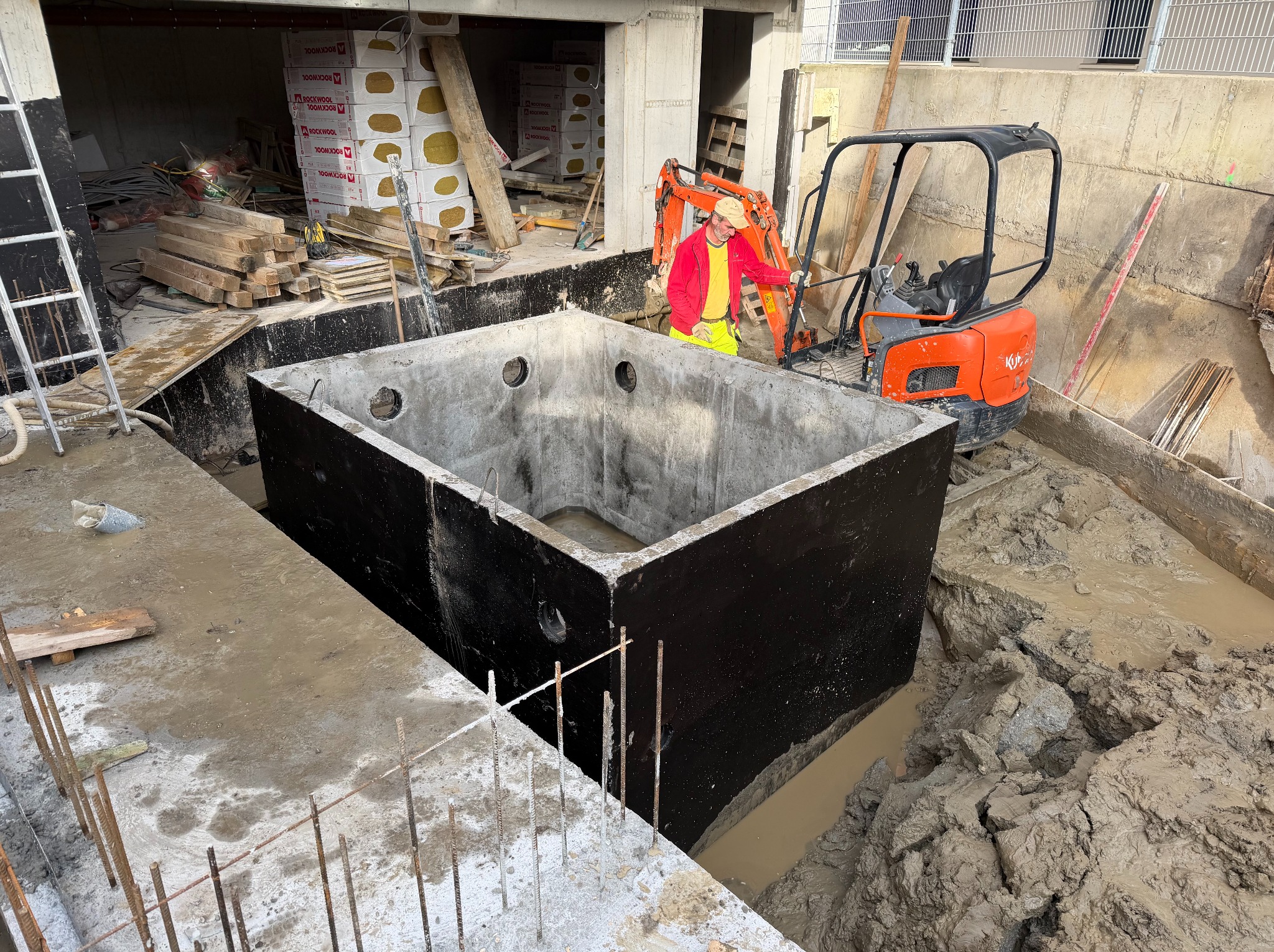 Betonowy zbiornik w wykopie, obok minikoparka i pracownik w odblaskowej odzieży. Widoczne materiały budowlane i elementy konstrukcyjne. Ujęcie z góry, dzień.