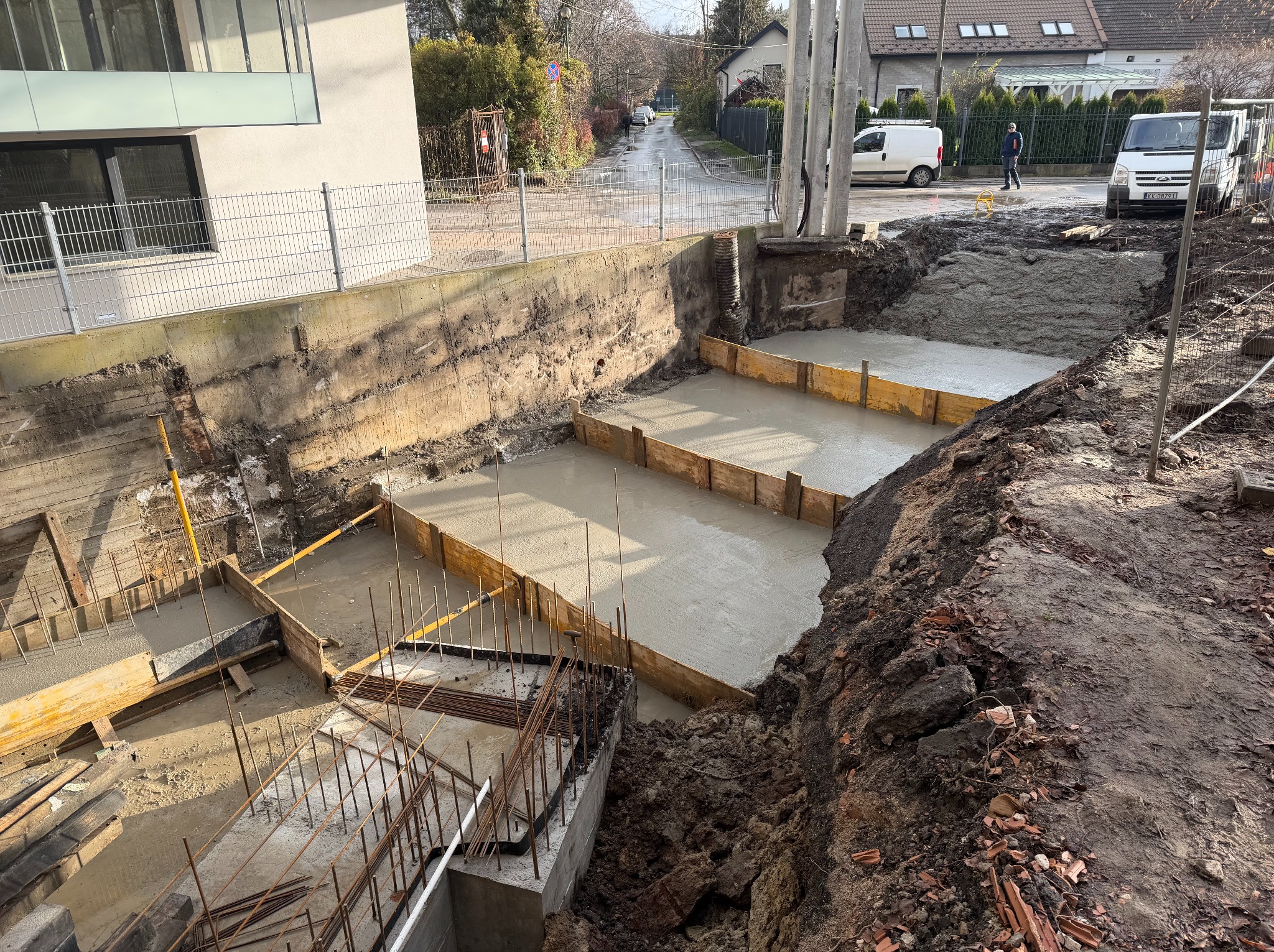 Wylany beton w szalunkach z widocznym zbrojeniem na budowie domu jednorodzinnego. Ujęcie z góry, w tle zabudowa mieszkalna i ulica.