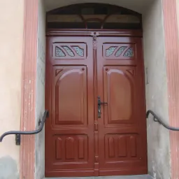 Lakierowane na brązowo dwuskrzydłowe drzwi wejściowe z przeszkleniami o nieregularnym kształcie, ozdobione metalowymi uchwytami po bokach, osadzone w ścianie z ozdobnymi pilastrami.