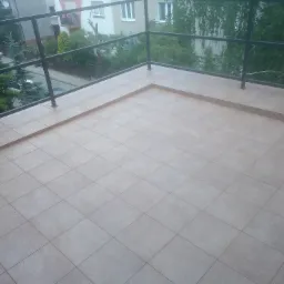 Wykafelkowany balkon z beżowymi płytkami, czarna metalowa balustrada, widok na zieleń i budynki w tle.