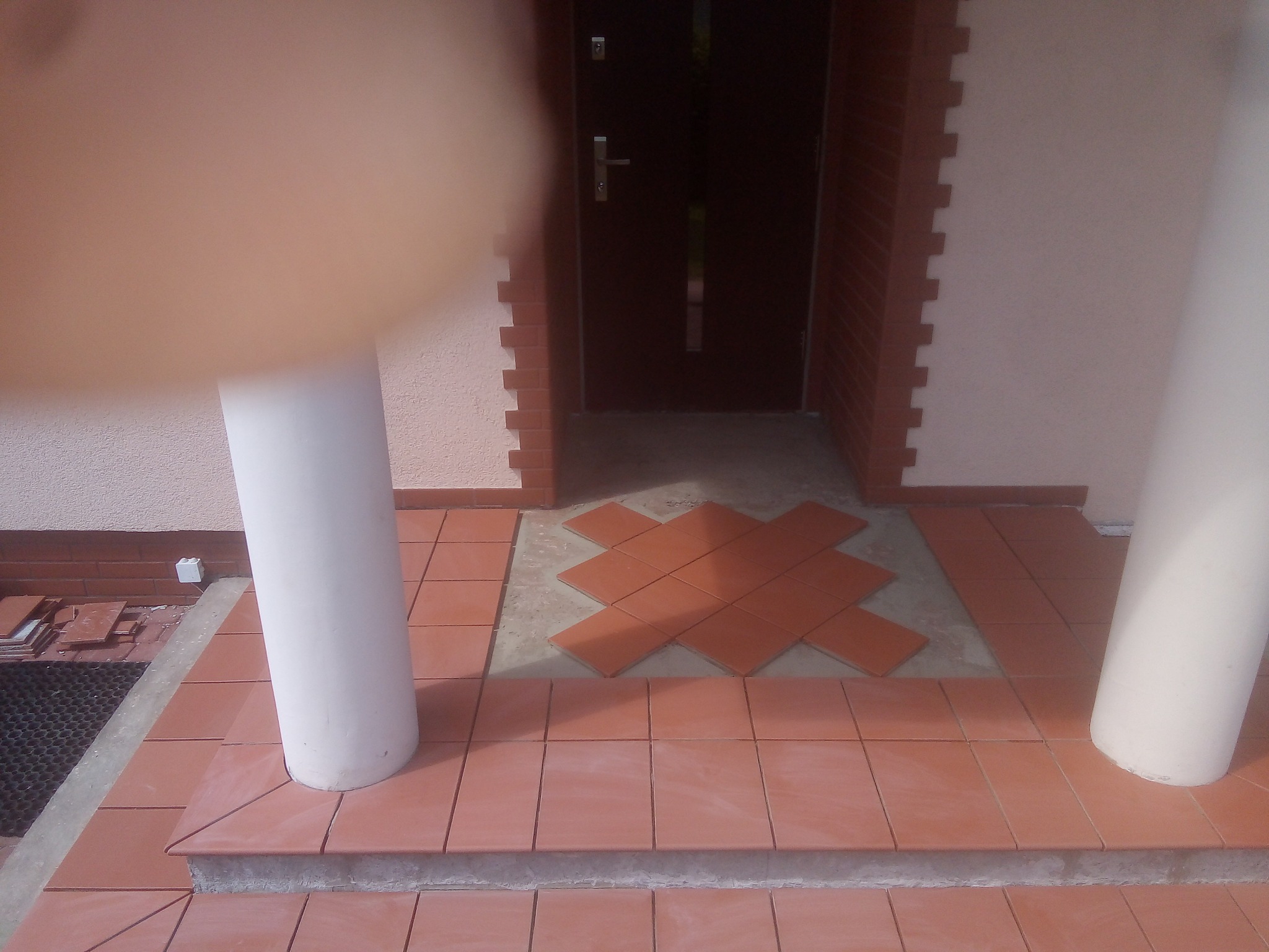 Układanie terakoty na schodach wejściowych, widoczne dwie białe kolumny, fragment dłoni w rogu kadru oraz wzór z płytek w kształcie krzyża na podeście.