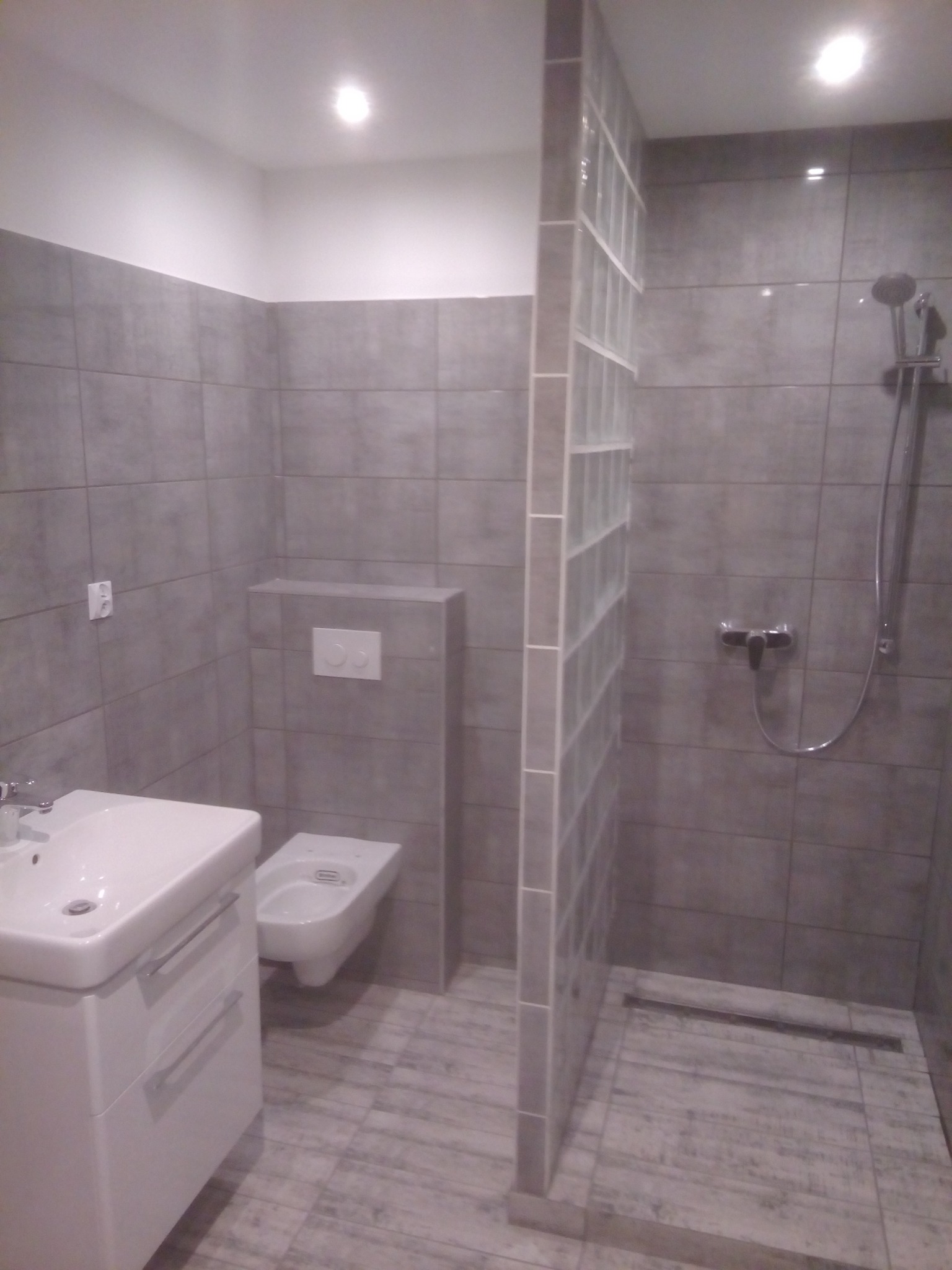 Szara łazienka z kabiną prysznicową oddzieloną szklanymi luksferami, umywalką z szafką, toaletą podwieszaną i szarą armaturą.