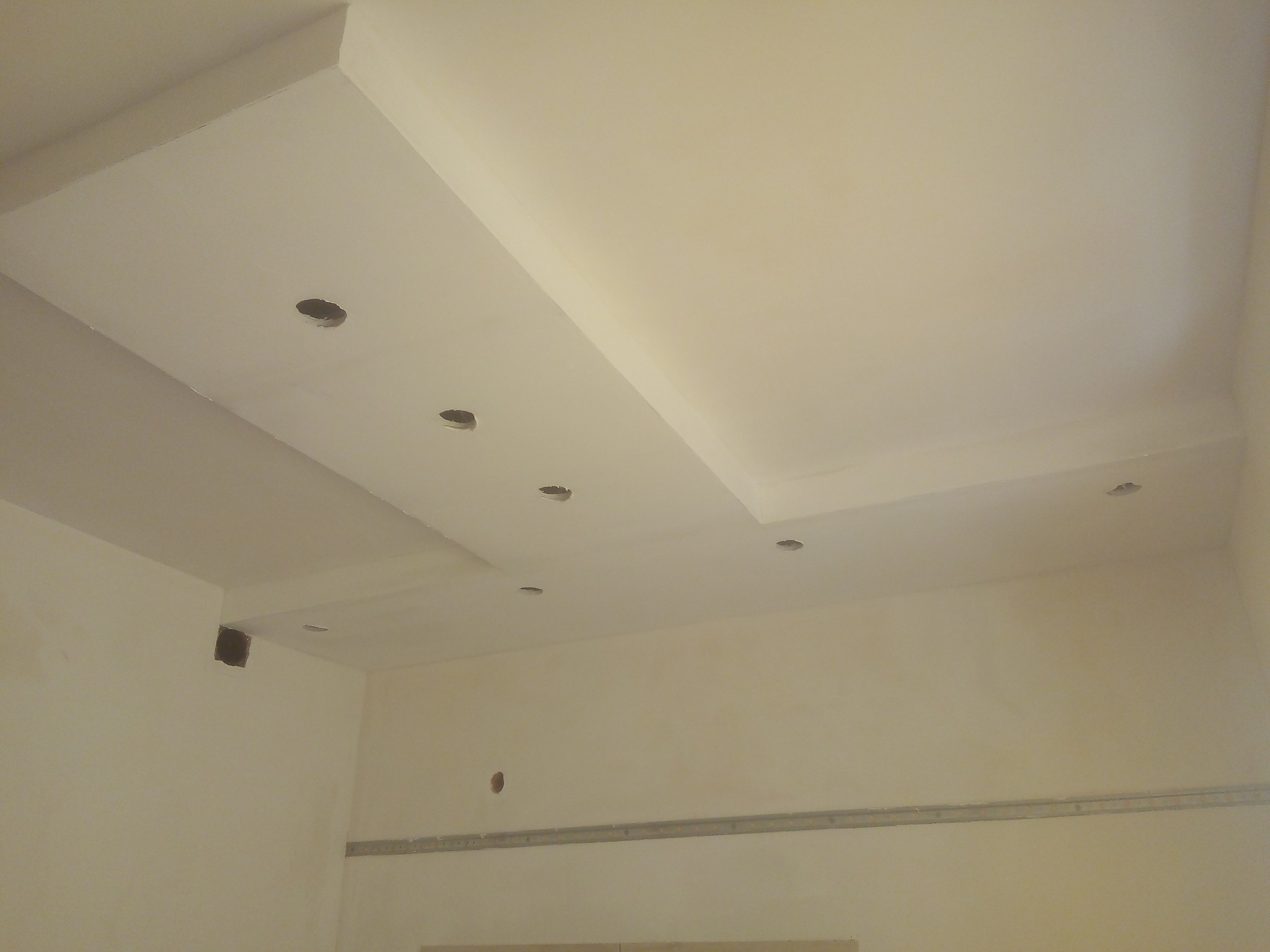 Wykończony sufit podwieszany z otworami na oświetlenie punktowe, świeżo pomalowany na kremowo, widoczne elementy montażowe na ścianach.