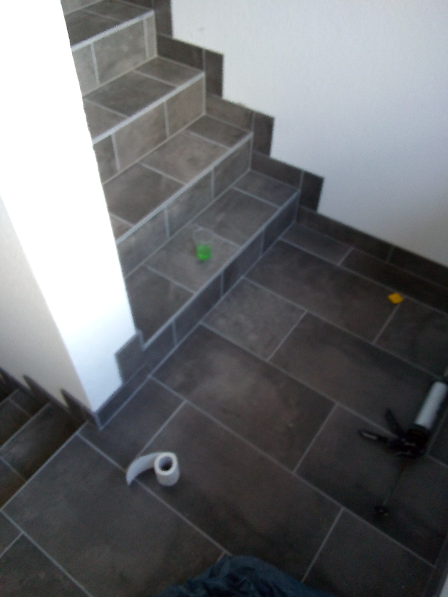 Ułożone płytki podłogowe i schodowe w odcieniach szarości z widoczną rolką taśmy malarskiej, zielonym kubkiem i pistoletem do silikonu, sugerujące etap prac wykończeniowych.