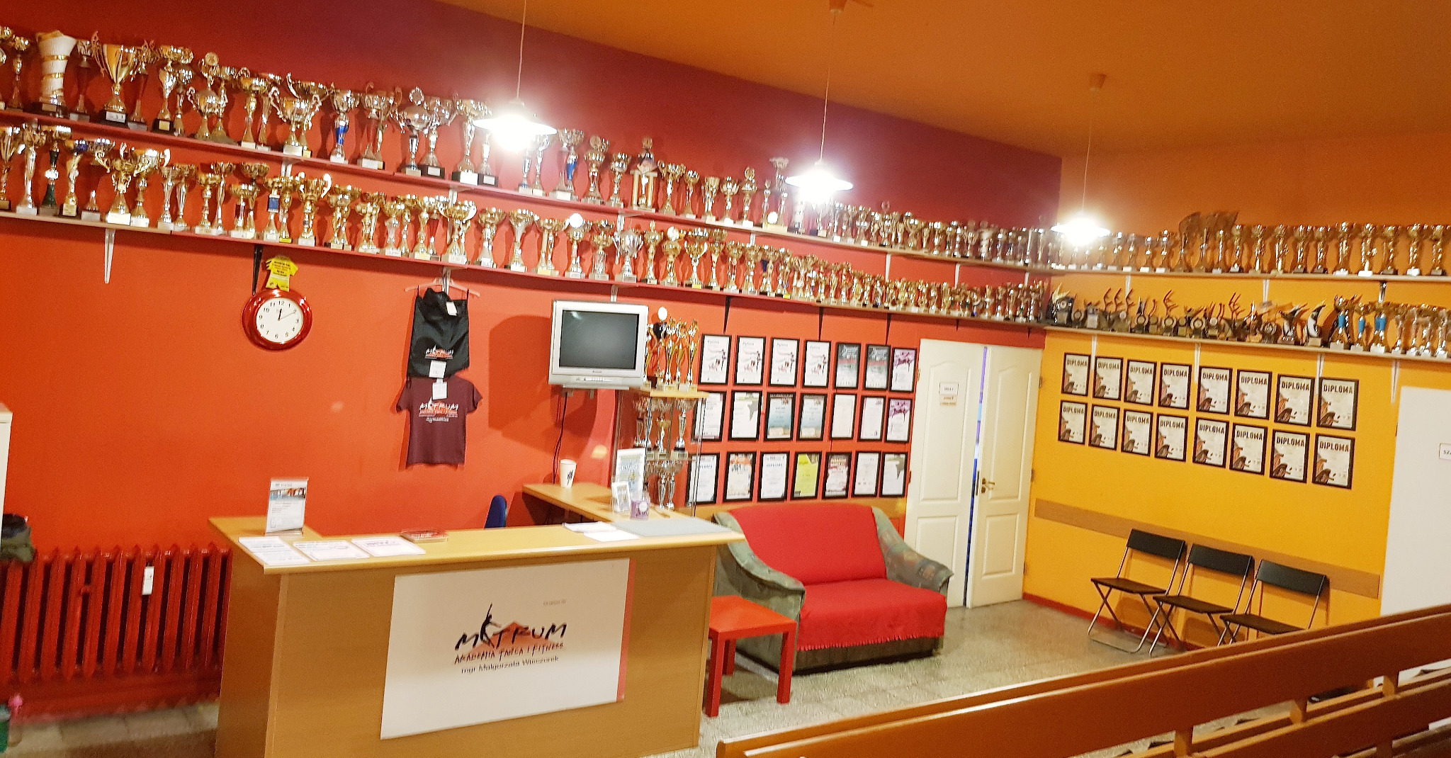 Wnętrze z recepcją, ścianami w odcieniach pomarańczy i żółci, udekorowanymi rzędami pucharów i dyplomów, z widocznym logo i nazwą Akademii Tańca i Fitness