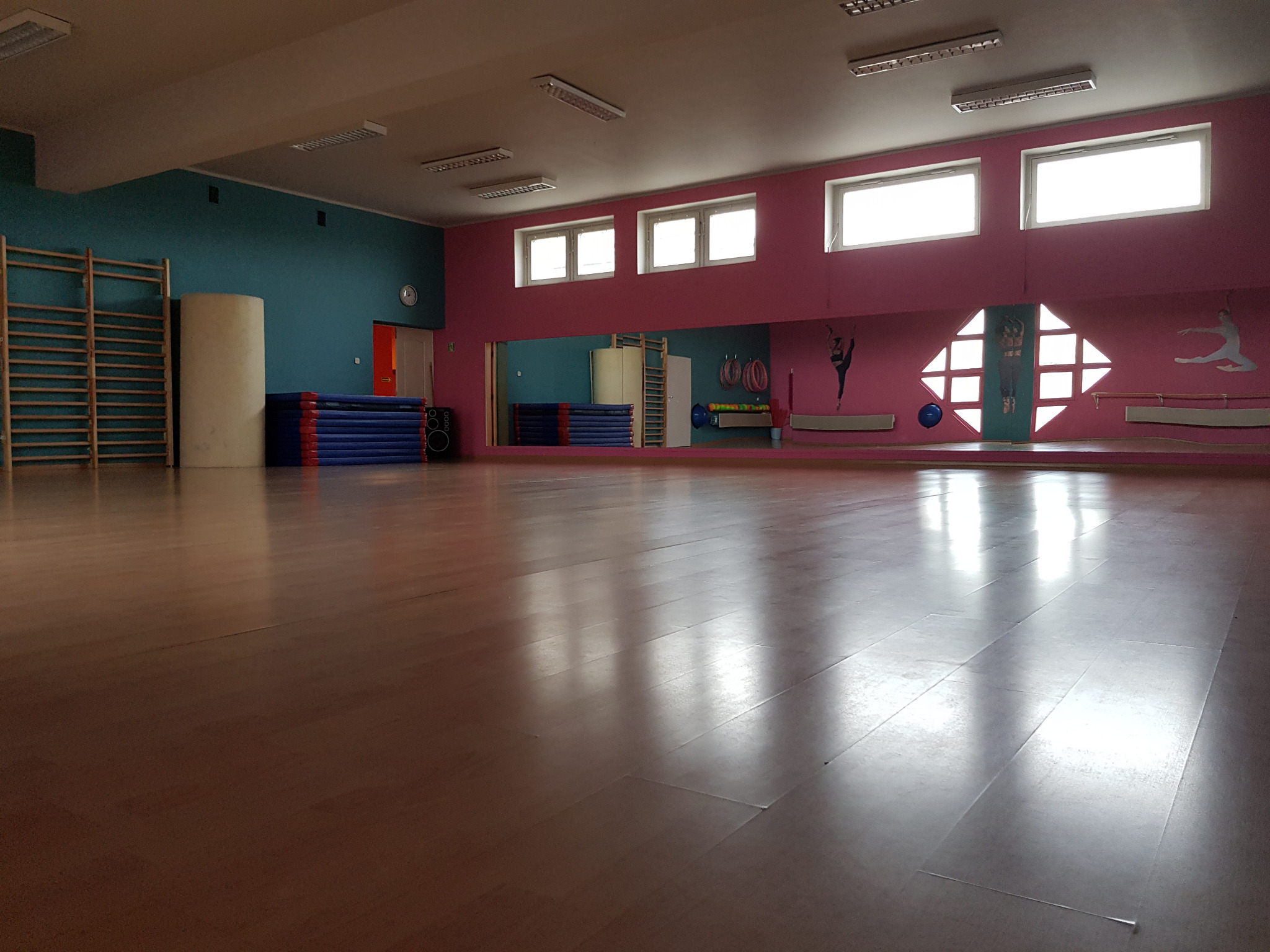 Pusta sala z drewnianą podłogą, drabinkami gimnastycznymi przy turkusowej ścianie, materacami i lustrem z naklejkami sylwetek tancerzy na różowej ścianie.
