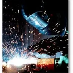 Intensywne iskry podczas spawania metalowych elementów, widoczna maska spawalnicza i fragment spawanej konstrukcji w słabym oświetleniu.