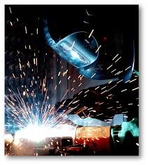 Intensywne iskry podczas spawania metalowych elementów, widoczna maska spawalnicza i fragment spawanej konstrukcji w słabym oświetleniu.