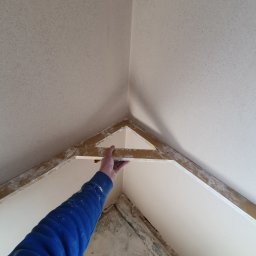 Dam-Bud - Ręka w niebieskiej bluzie przytrzymuje łatę tynkarską w rogu pomieszczenia, widoczne świeżo nałożone tynki na ścianach i suficie, zarysowana drewniana konstrukcja w tle.