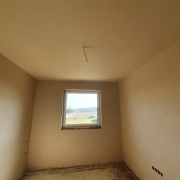 Dam-Bud - Wnętrze pokoju po nałożeniu gładzi gipsowej na ściany i sufit, widoczne przygotowane otwory pod gniazdka elektryczne i wystający kabel z sufitu, okno z widokiem na pole.