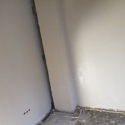 Dam-Bud - Świeżo wykończony róg pokoju z gładkimi, białymi ścianami, widoczne instalacje elektryczne w ścianie i rurki przy podłodze, surowy beton na podłodze.