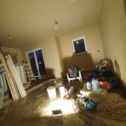 Dam-Bud - Wnętrze w trakcie prac wykończeniowych: świeżo otynkowane ściany i sufit w odcieniach beżu, widoczne narzędzia, worki z materiałami budowlanymi, drabina, okno i drzwi balkonowe, tymczasowe...