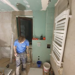 Dam-Bud - Remont łazienki w trakcie: instalacja grzejnika naściennego, płyty gipsowo-kartonowe przygotowane pod wykończenie, widoczny pracownik z miarką, płytki podłogowe i narzędzia.