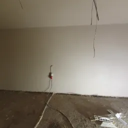 Wnętrze pokoju po nałożeniu gładzi, widoczne wystające kable, gniazdko elektryczne oraz narzędzia na podłodze.