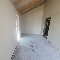 Dam-Bud - Wnętrze pokoju po nałożeniu gładzi, widoczne ślady po pracy na podłodze, rękawice robocze i narzędzia malarskie, surowe drewniane belki sufitowe, przygotowane do dalszych prac wykończeniowych.