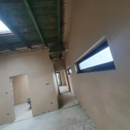 Surowe wnętrze domu po nałożeniu tynków, widoczne okno dachowe, okno boczne i otwory drzwiowe, podłoga zabezpieczona folią.