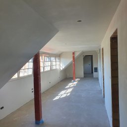 Dam-Bud - Poddasze w trakcie remontu: ściany i sufit pokryte świeżym, jasnym tynkiem, widoczne ślady po gładzi, drewniane słupy zabezpieczone folią, okna dachowe wpuszczające ostre promienie słoneczne.