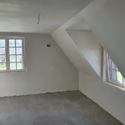 Poddasze po nałożeniu gładzi, widoczne dwa okna o różnej wielkości, ściany przygotowane do dalszych prac wykończeniowych, wystające kable ze ściany i sufitu.