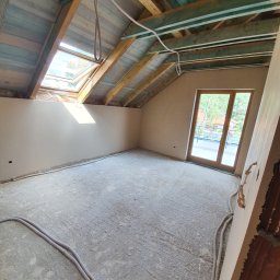 Dam-Bud - Poddasze w trakcie prac wykończeniowych: ściany pokryte świeżym tynkiem, widoczne okno dachowe, drewniana konstrukcja dachu i kable elektryczne na podłodze.