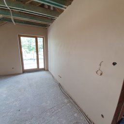 Dam-Bud - Poddasze po nałożeniu gładzi, widoczne drewniane belki stropowe, okno balkonowe, kable elektryczne w ścianie oraz na podłodze pokrytej resztkami gładzi.