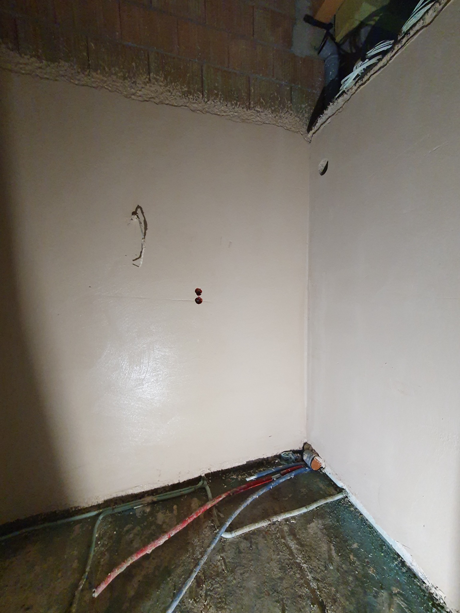 Świeżo nałożony, gładki tynk w rogu pomieszczenia, z widocznymi przewodami instalacji elektrycznej i hydraulicznej przy podłodze, przygotowany do dalszych prac wykończeniowych.