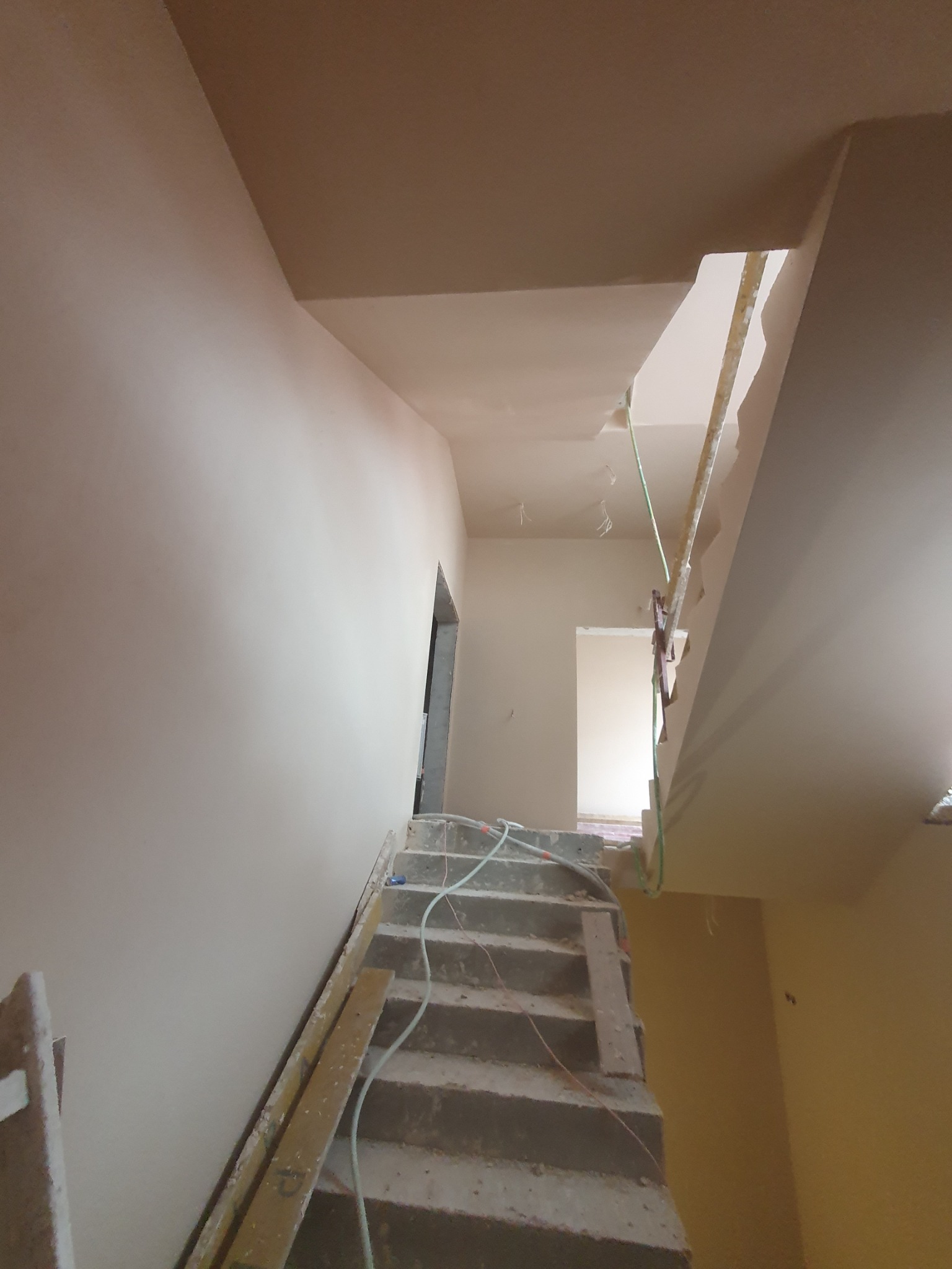 Wnętrze domu w trakcie prac wykończeniowych: świeżo otynkowane ściany i sufit w odcieniach beżu, schody betonowe z widocznymi przewodami instalacji, przygotowane do dalszych prac.