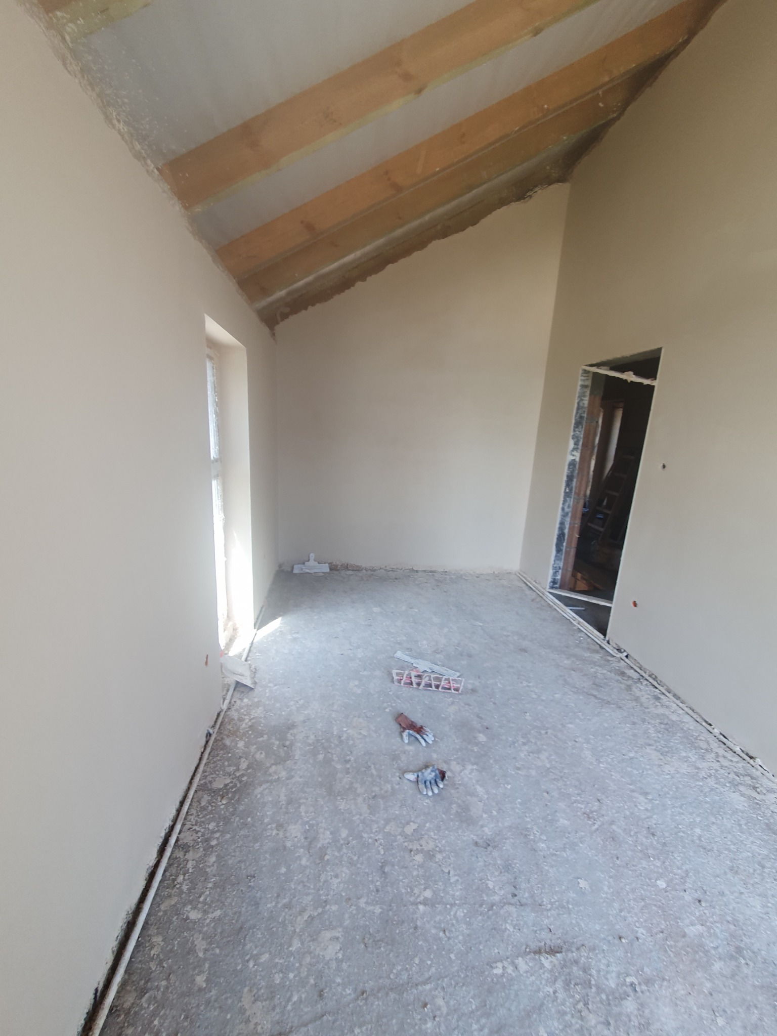 Wnętrze pokoju po nałożeniu gładzi, widoczne ślady po pracy na podłodze, rękawice robocze i narzędzia malarskie, surowe drewniane belki sufitowe, przygotowane do dalszych prac wykończeniowych.