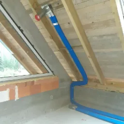 Montaż elastycznych, niebieskich przewodów w systemie wentylacyjnym poddasza, widoczne elementy mocujące i fragment okna dachowego.