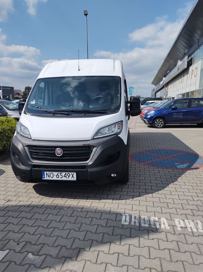 Biały samochód dostawczy Fiat Ducato z numerem rejestracyjnym NO 6549X, zaparkowany na parkingu z kostki brukowej przed salonem samochodowym w słoneczny dzień.