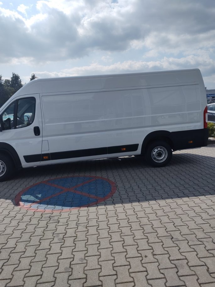 Biały furgon dostawczy zaparkowany na brukowanym parkingu z niebieskim znakiem zakazu parkowania na ziemi.