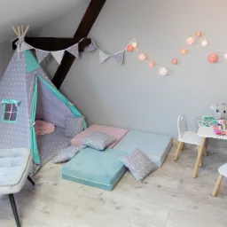 Pokój zabaw dla dziecka z tipi w gwiazdki, stolikiem z krzesełkami i kolorowymi poduszkami pod skosem dachu.