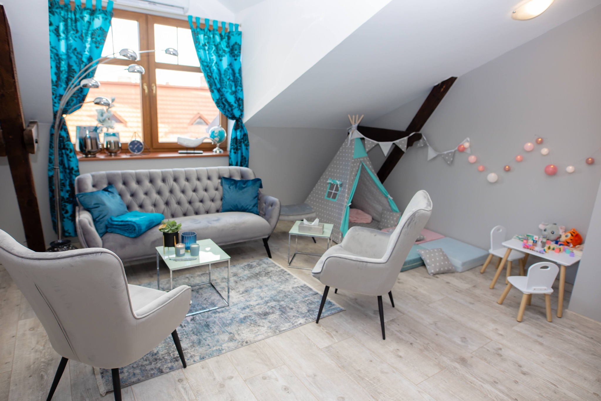 Jasne, przytulne wnętrze gabinetu psychologicznego z szarym kanapą, turkusowymi akcentami, dziecięcym tipi i stolikiem z zabawkami.