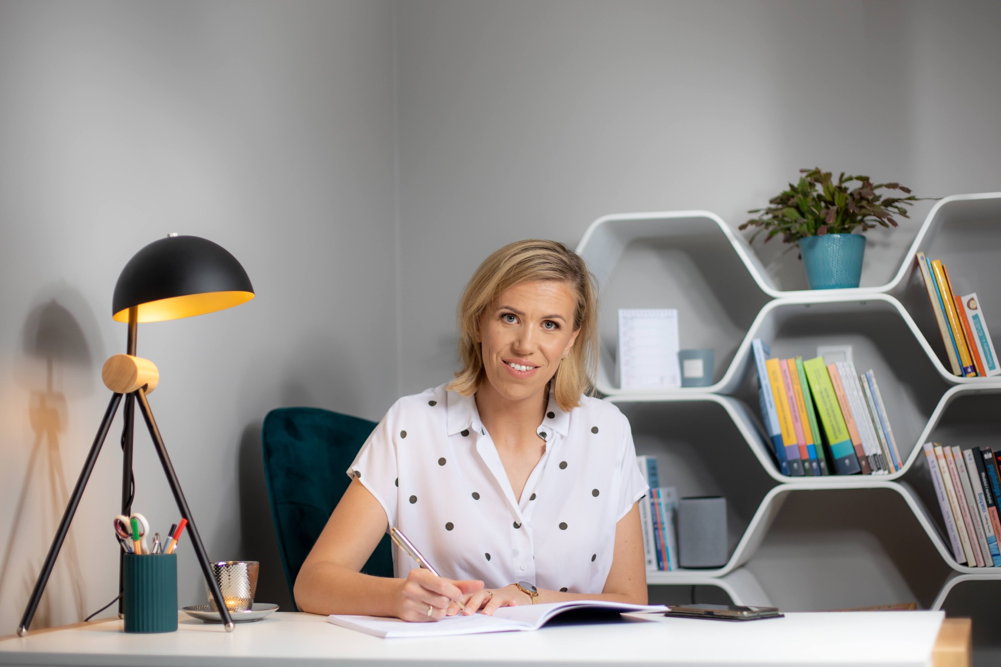 Kobieta w białej koszuli w czarne kropki pisze długopisem w notatniku na białym biurku, w tle półka z książkami i lampa na trójnogu.