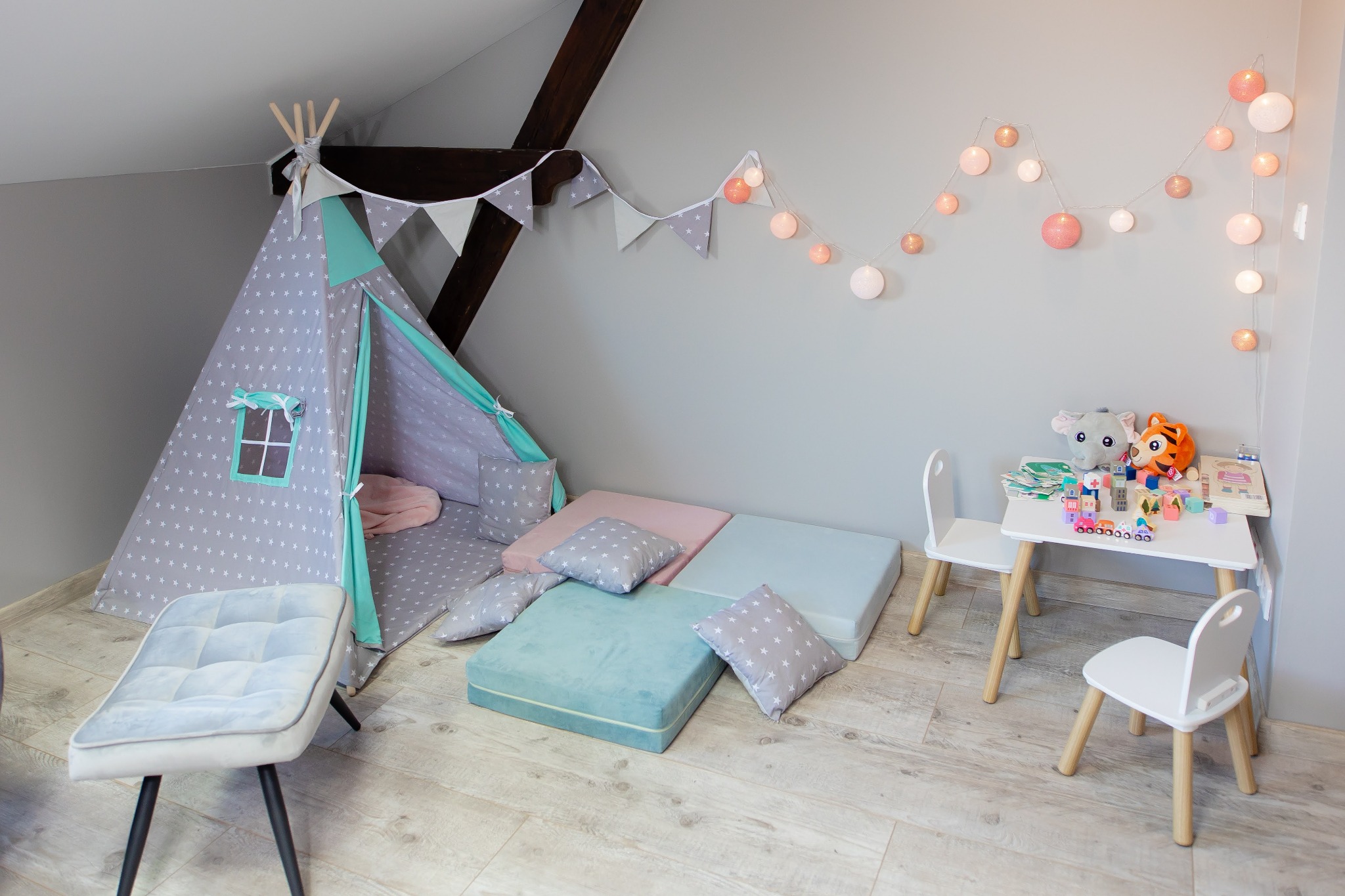 Pokój zabaw dla dziecka z tipi w gwiazdki, stolikiem z krzesełkami i kolorowymi poduszkami pod skosem dachu.