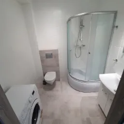 Jasna łazienka po remoncie z kabiną prysznicową, umywalką z szafką, toaletą podwieszaną i pralką Indesit.