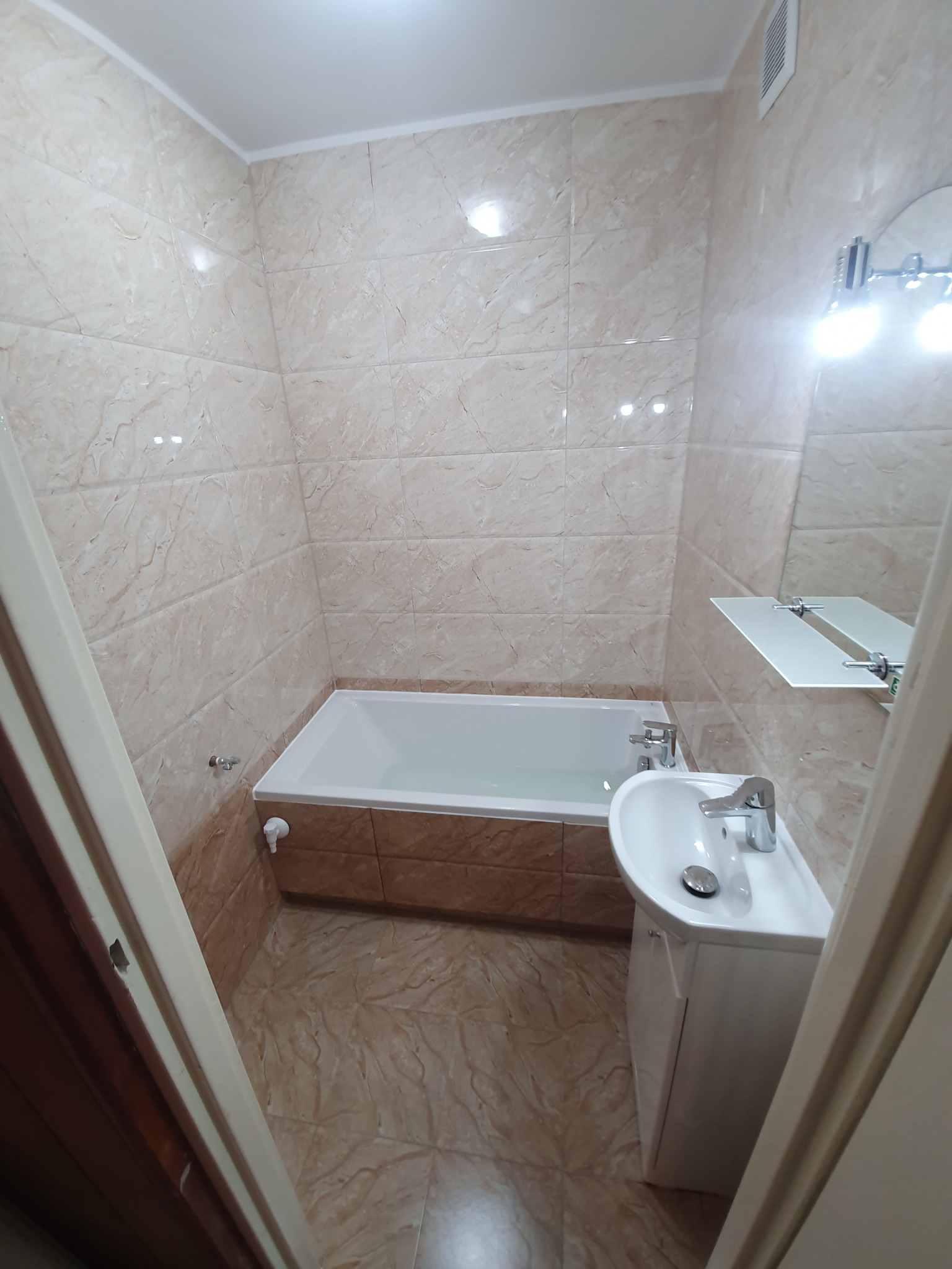 Wykończona łazienka z beżowymi płytkami imitującymi marmur, biała wanna wypełniona wodą, umywalka z szafką i lustro z oświetleniem, widok zza framugi drzwi.