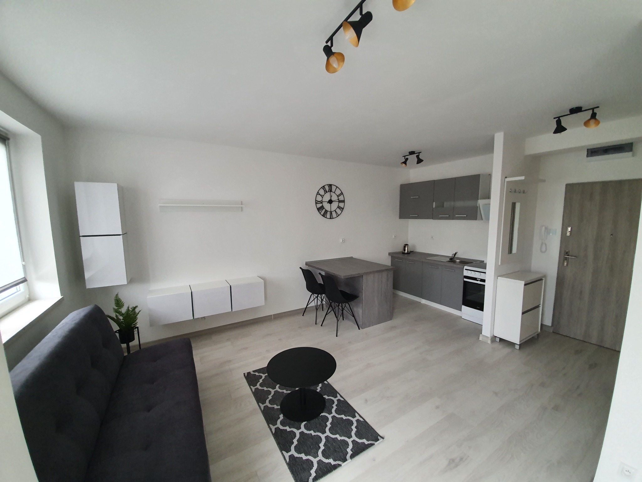 Nowoczesne wnętrze mieszkania po remoncie: szara sofa, białe szafki RTV, okrągły stolik kawowy na wzorzystym dywanie, aneks kuchenny z szarymi szafkami i wyspą, zegar na ścianie, oświetlenie punktowe.